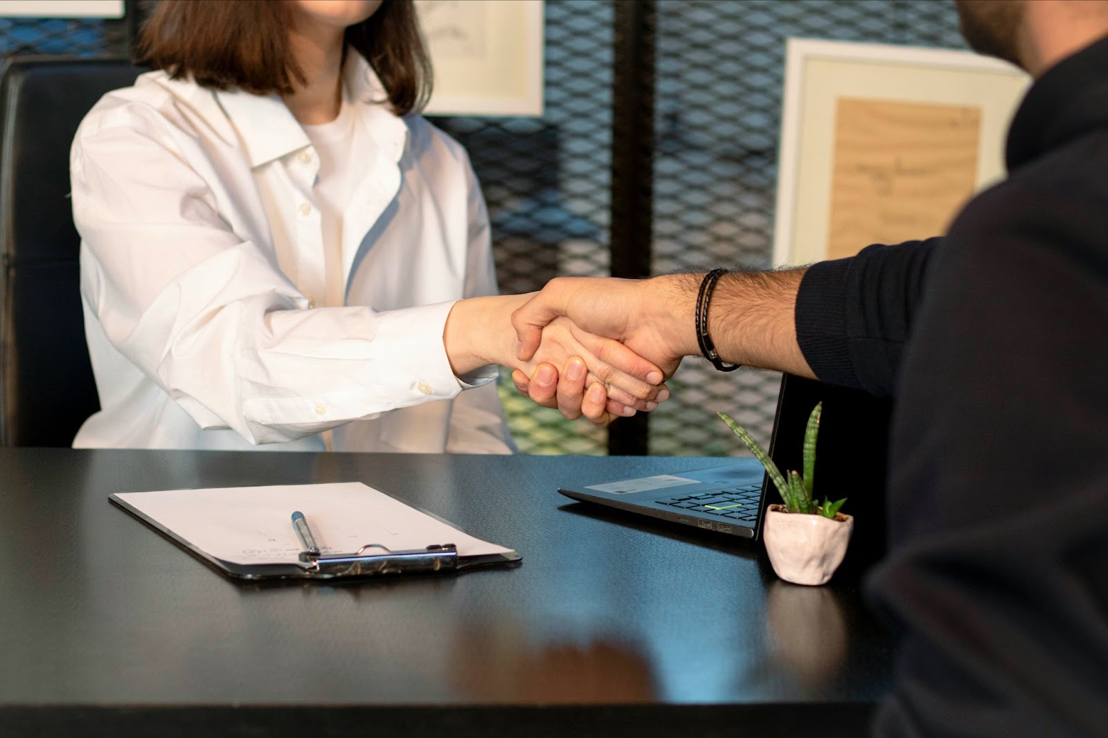 Two HR leaders shaking hands over a desk with a clipboard and a laptop.